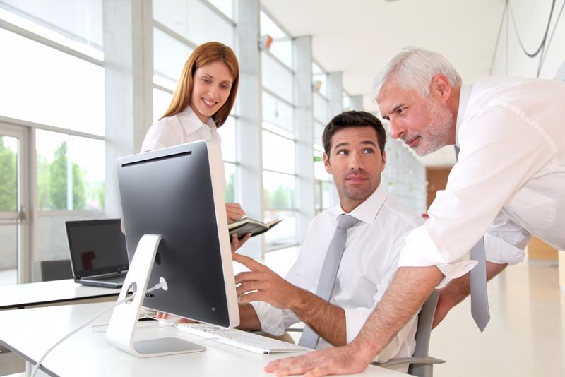 An older worker helps two young workers by a computer in an office