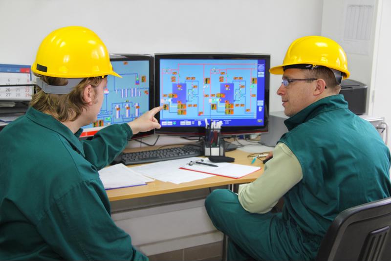Two industrial workers in control room communicating around desk with monitors. 