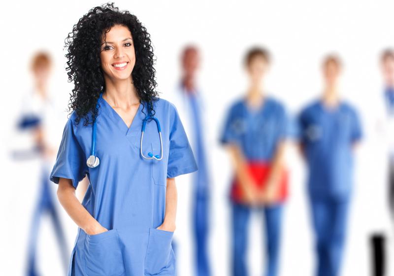 Woman wearing blue scrubs and stethoscope, with other health care workers standing behind her in the background. 