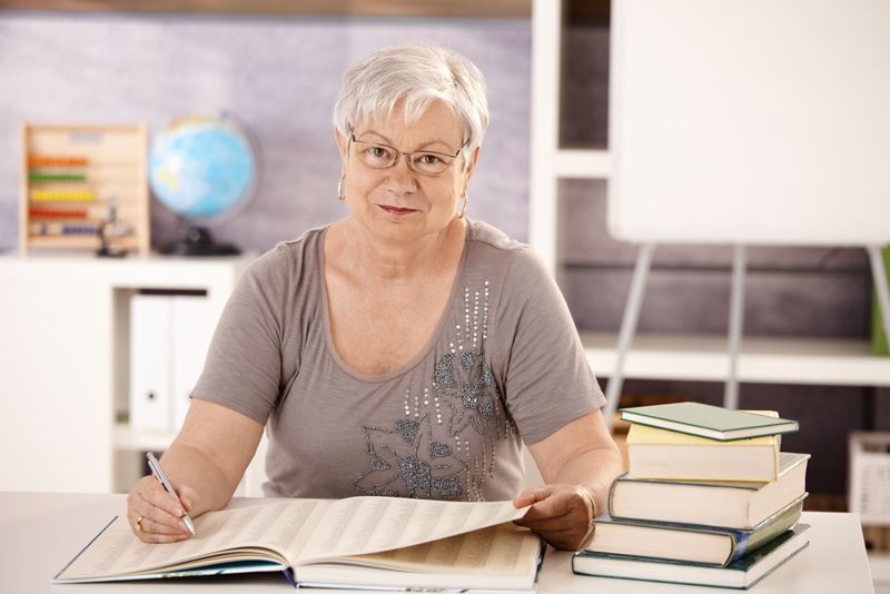 Older woman sitting at a desk with a stack of books in a classroom. 
