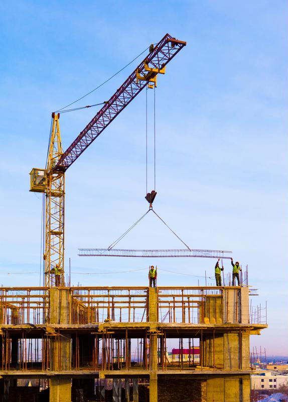Workers guiding beam lowered by crane on building under construction.