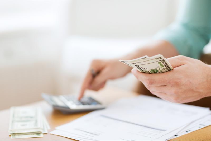 A man sitting at a table with a stack of invoices and cash while using a calculator and holding cash in one hand. 