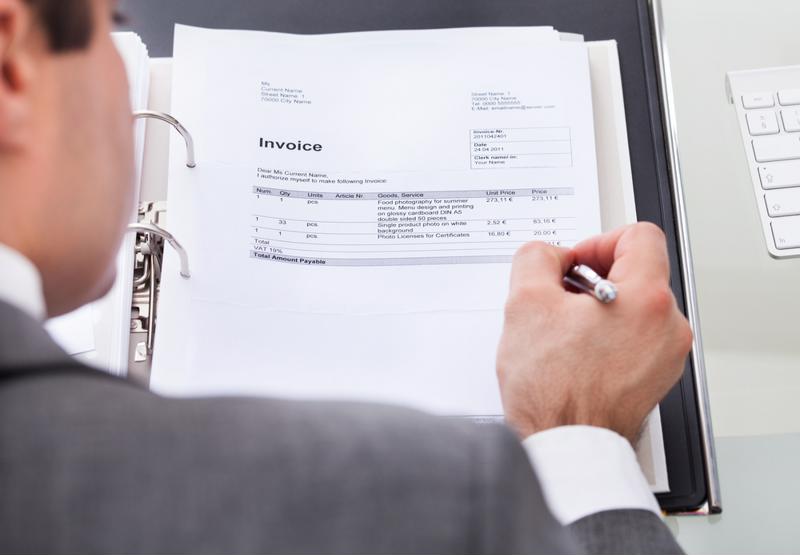 Over the shoulder view of a business man in a suit filling out paper invoice in a binder with silver pen. 