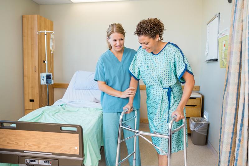 Nurse holding the arm of a young female hospital patient wearing hospital gown and using a walker. 