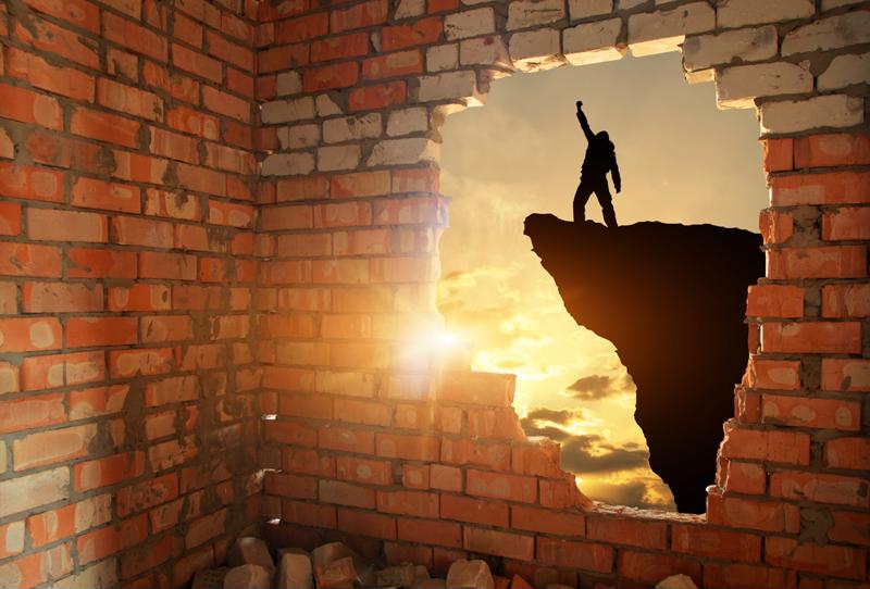 Hole in a brick wall reveals shadow silhouette of a person with their fist raised victoriously in the air, standing on the edge of a cliff. 