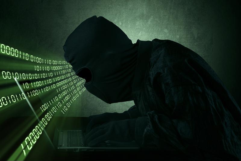 Man in all black - gloves, mask, jacket - typing on a laptop with binary code coming out of his face and heading into the laptop screen. 