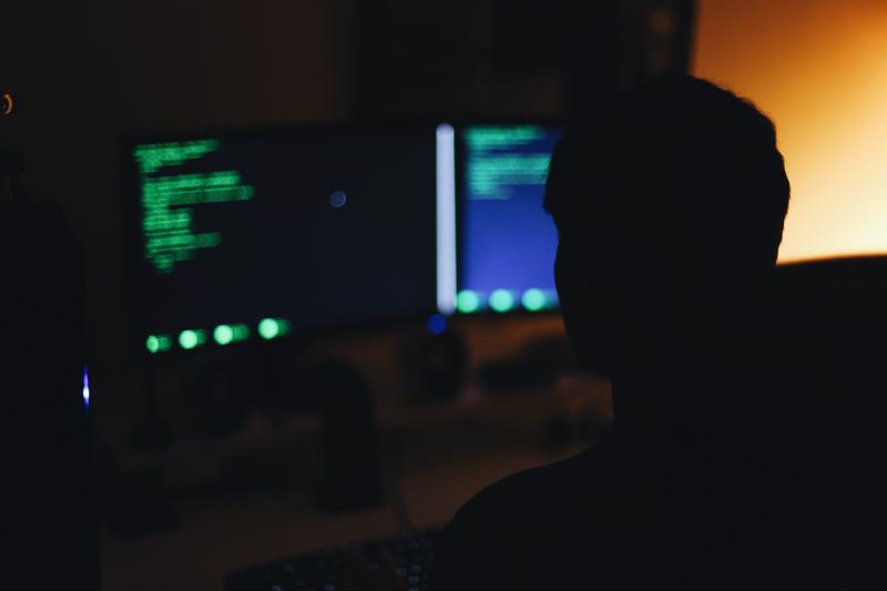 Hacker in darkened room in front of computer workstation with dual monitors 