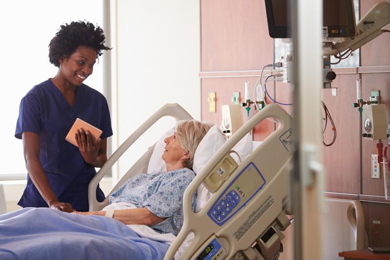 African American nurse wearing blue scrubs standing at the bedside of an elderly, female patient in a hospital room. 