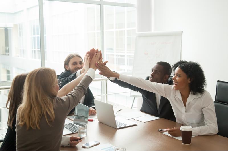Five office workers sitting around a conference room table, all reaching towards the middle for a group high-five.