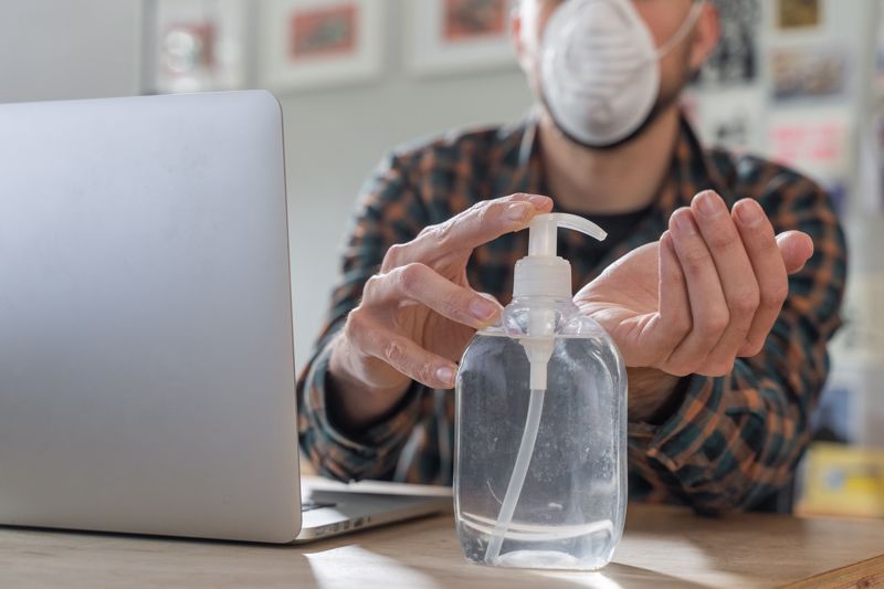 MAN WORKING FROM HOME WITH MASK AND HAND SANITIZER, SITTING IN FRONT OF LAPTOP AND PLACING SANITIZER IN OPEN HAND