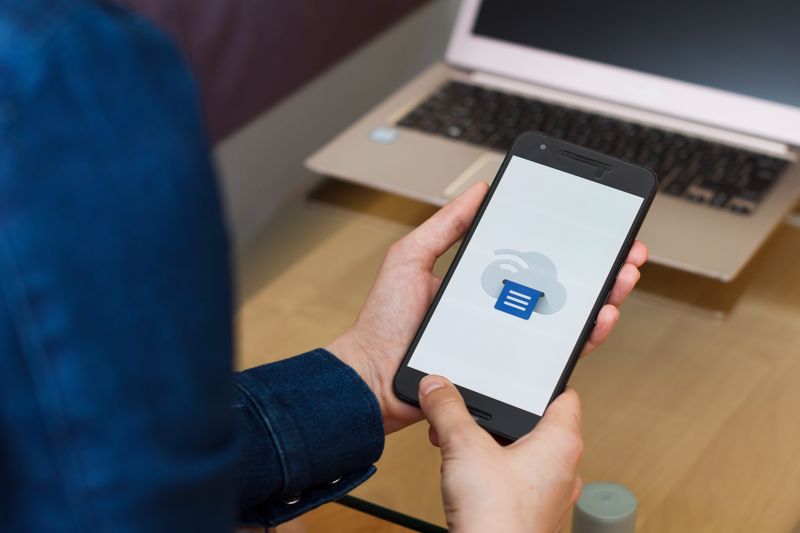 A person holding a smartphone with a print icon on the screen next to a laptop.