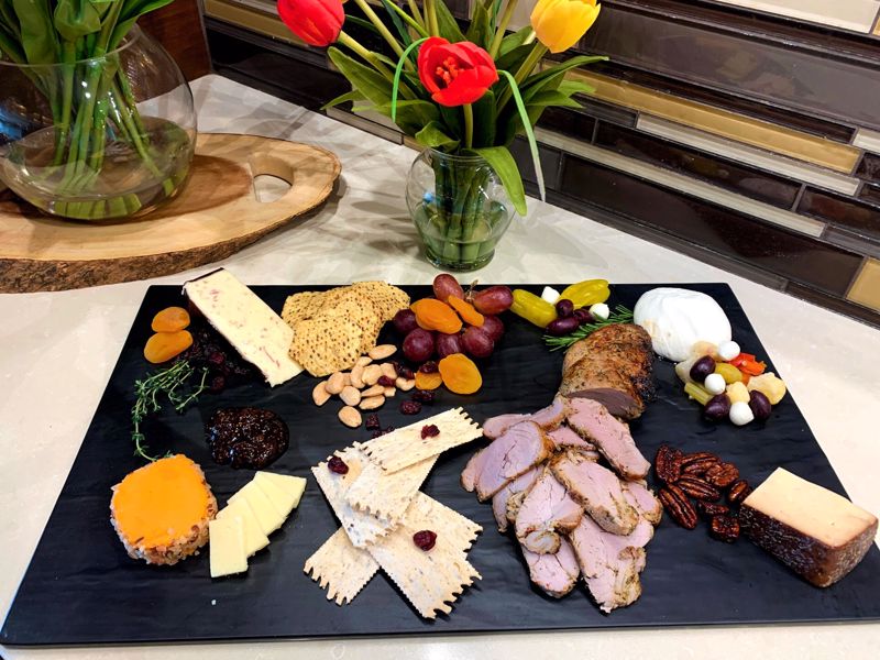 A charcuterie board of meat, cheese, grapes and nuts sits on a table with flowers in the background
