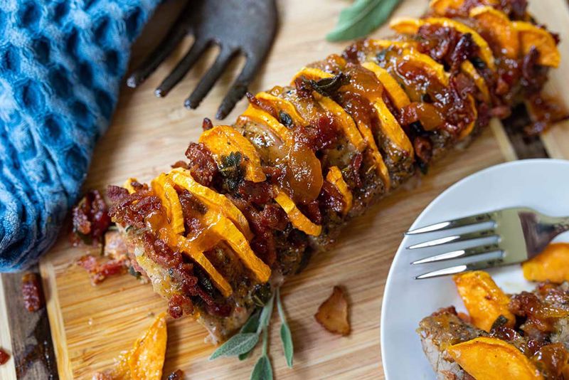 A Sage and Sweet Potato Hasselback Pork Tenderloin sits next to silverware on the table