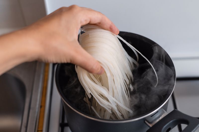 A person cooks rice noodles in a pot on the stove