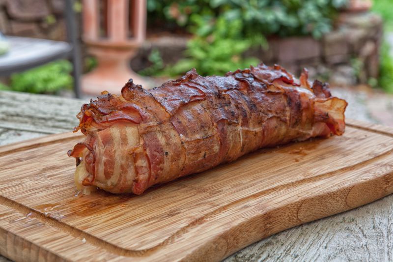Grilled bacon-wrapped  pork tenderloin sits on a wooden board.