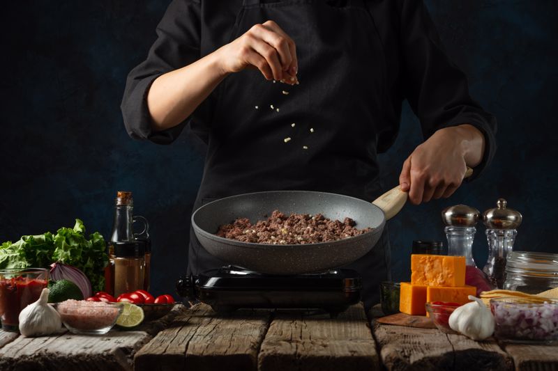 A chef prepares meat and spices in a skillet/pan