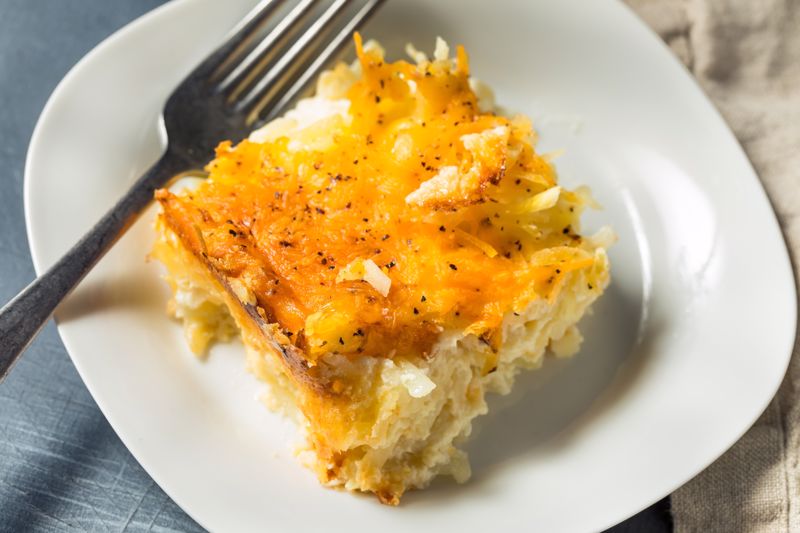 A slice of ham and hashbrown casserole sits on a plate next to a fork.